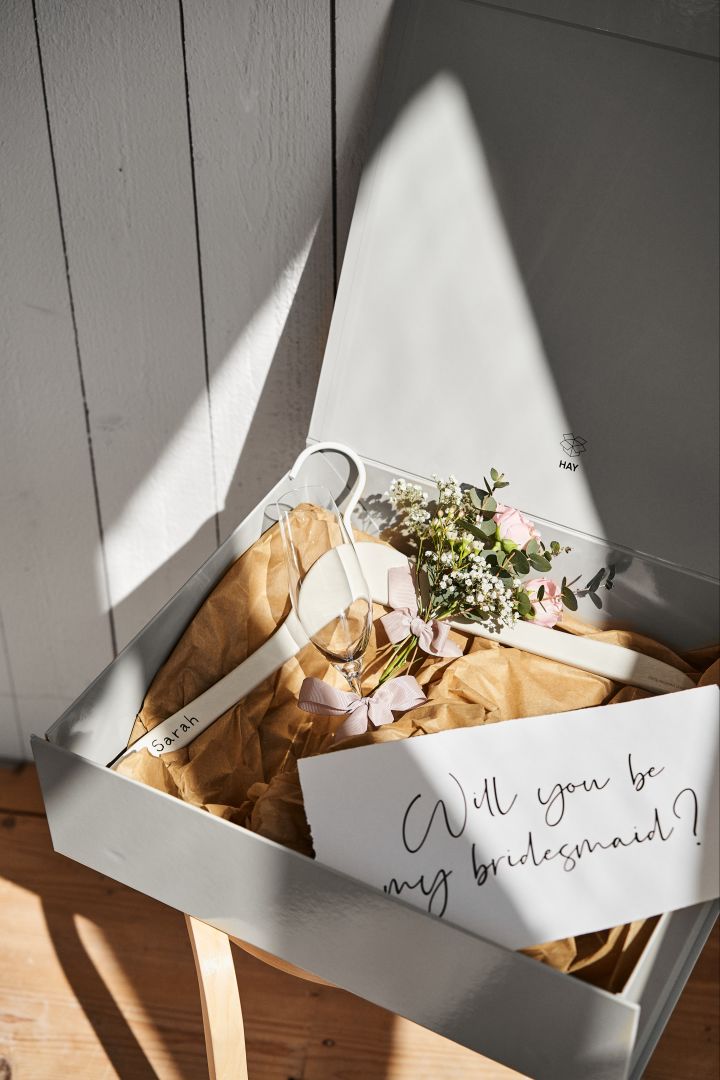 Verras je maid of honour met een cadeaubox. Hier zie je een cadeubox met een gepersonalisserde kledinghanger, champagneglas en bosje bloemen ingezoomd.