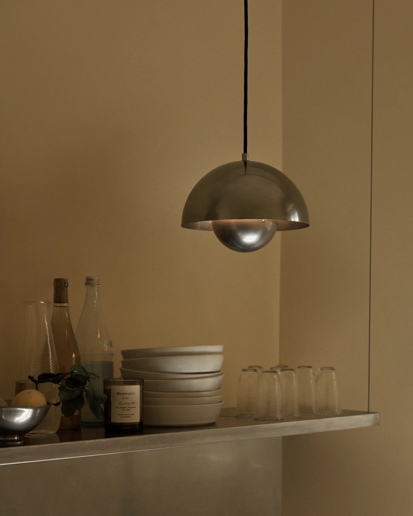 Een geborstelde metalen koepelhanglamp hangt boven een plank met kommen, flessen en glazen.