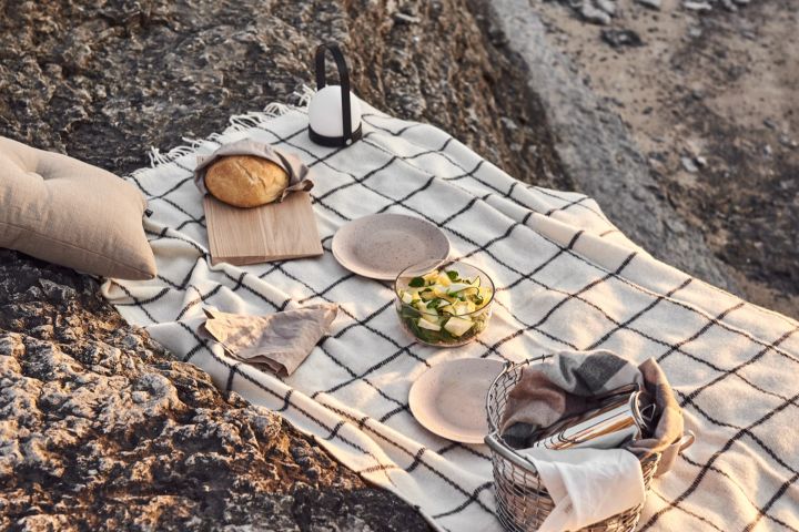 Geniet van een picknick op het strand bij zonsondergang met verschillende snacks, een deken en draagbare verlichting - hier de Carrie lamp in zwart van Audo Copenhagen. 