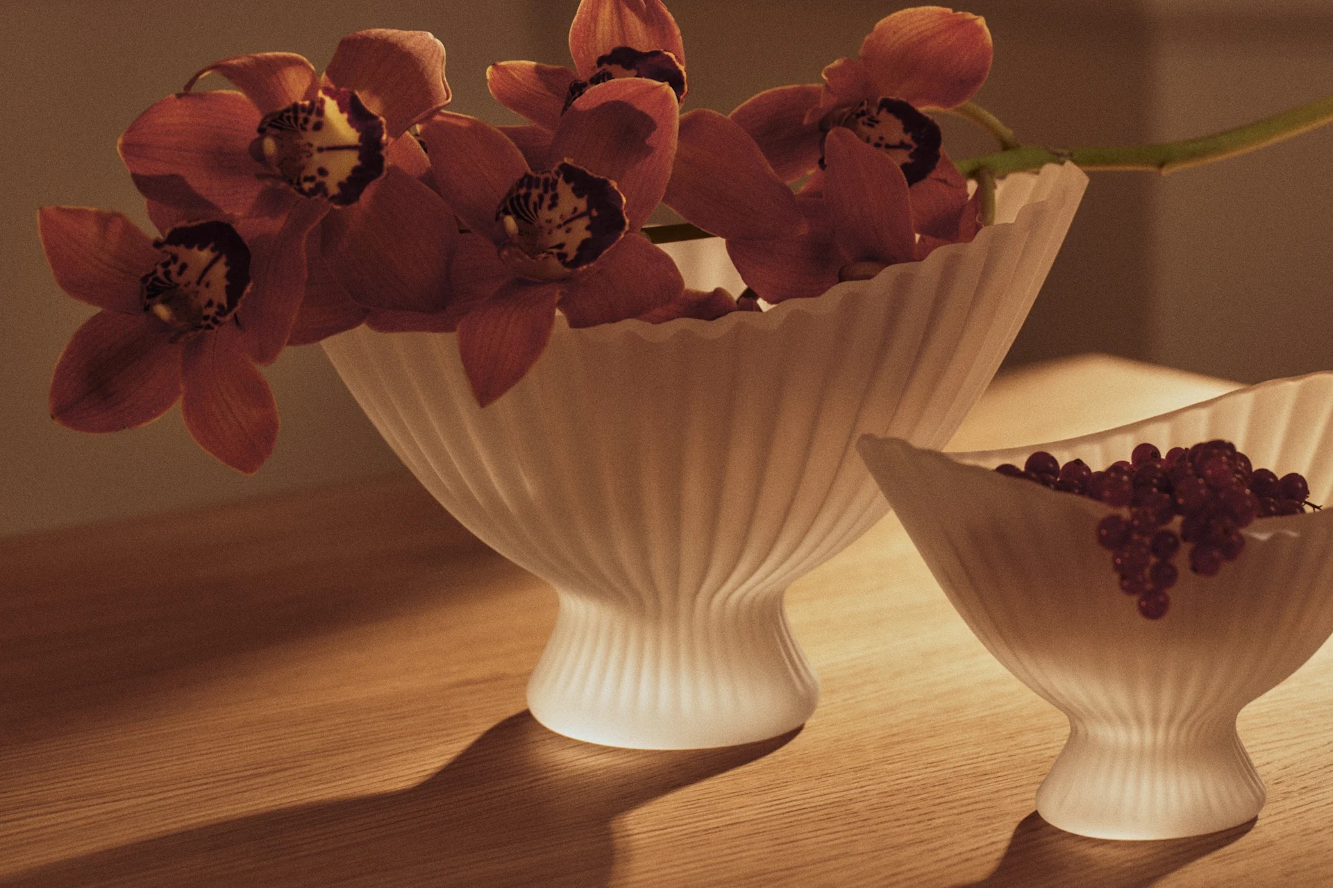 Rood-oranje orchideeën in geribbelde vaas en rode bessen in kleinere bijpassende schaal op houten tafel.