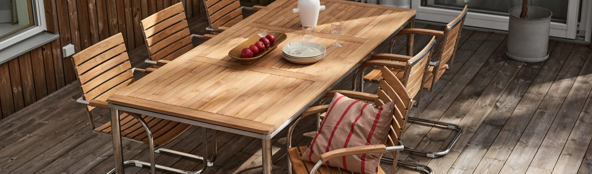Houten eettafel voor buiten met meerdere stoelen op een terras. De tafel heeft appels, borden en glazen. Een stoel heeft een gestreept kussen.