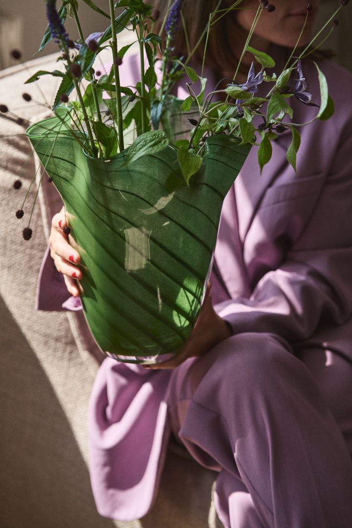 Een persoon in paarse kleding die de groene glasvaas Tiggy van Byon vasthoudt, een vaas met een geplooide, klokvormige uitstraling in erwt groen met donkere strepen.