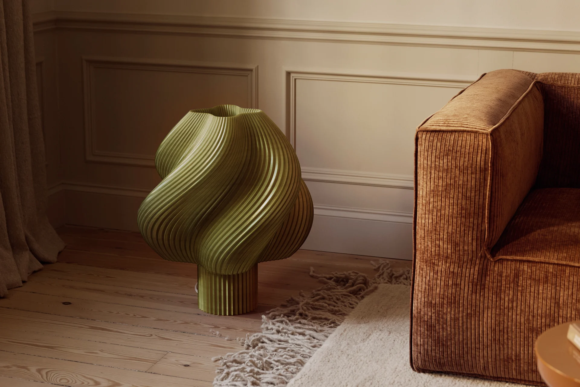 Een olijfgroen, sculpturaal, geribbeld object op een houten vloer naast een bruine corduroy bank en een getextureerd vloerkleed in een kamer met gegoten wanden.