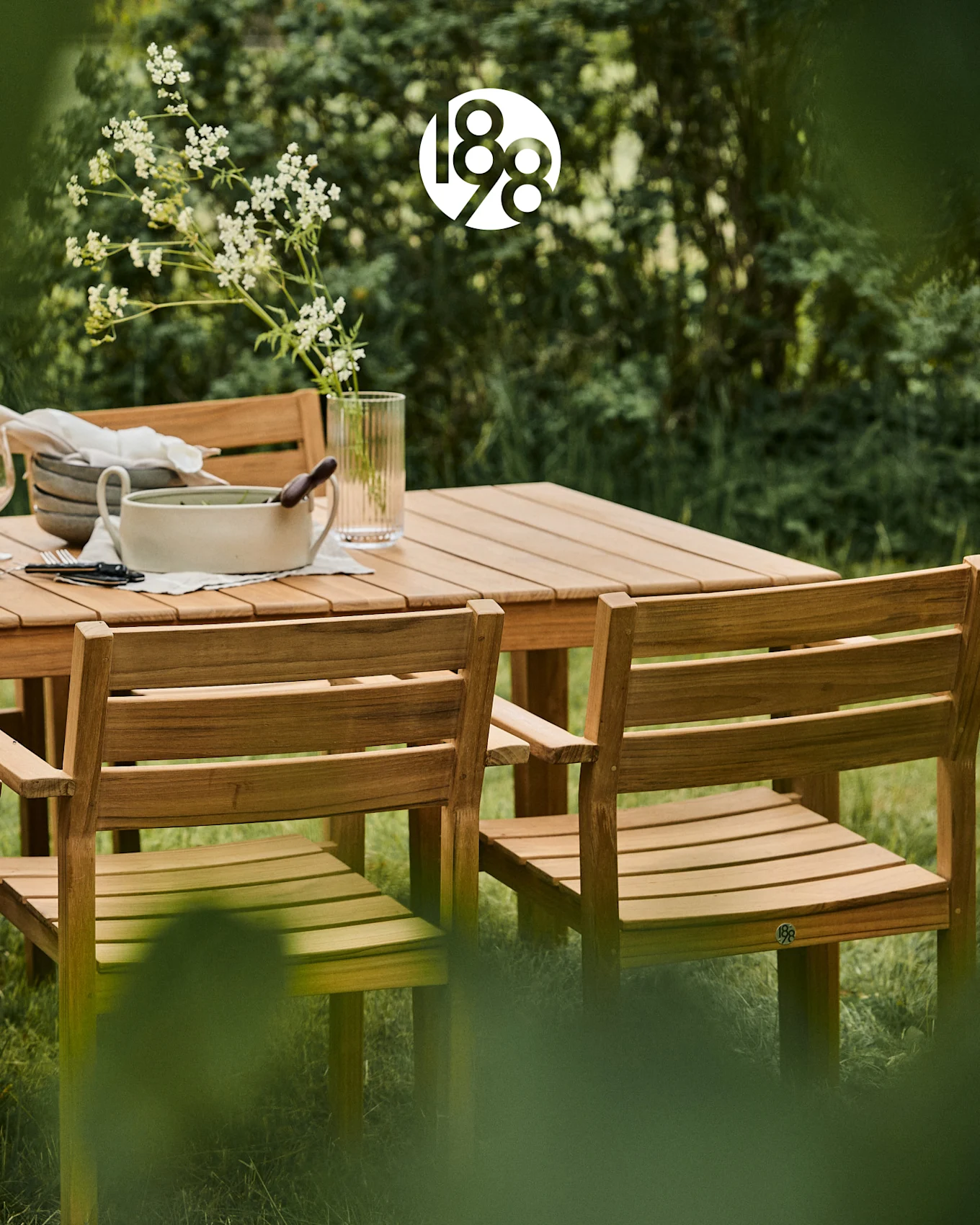 Houten buiten eetset met een tafel en stoelen in een weelderige groene tuin, met witte bloemen en serveerschalen.