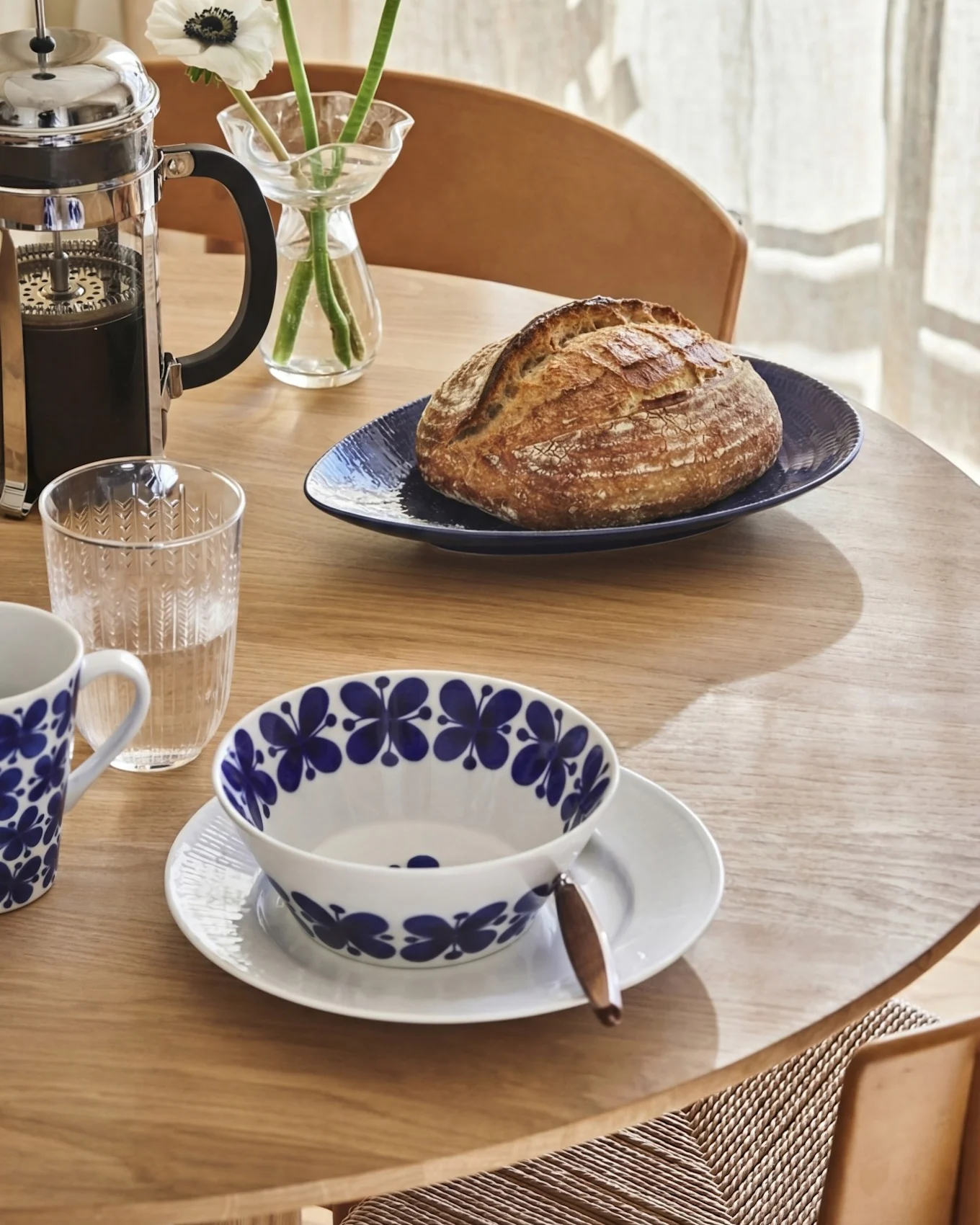 Ontbijttafel met French press, ambachtelijk brood, waterglas en kom en mok met patroon.