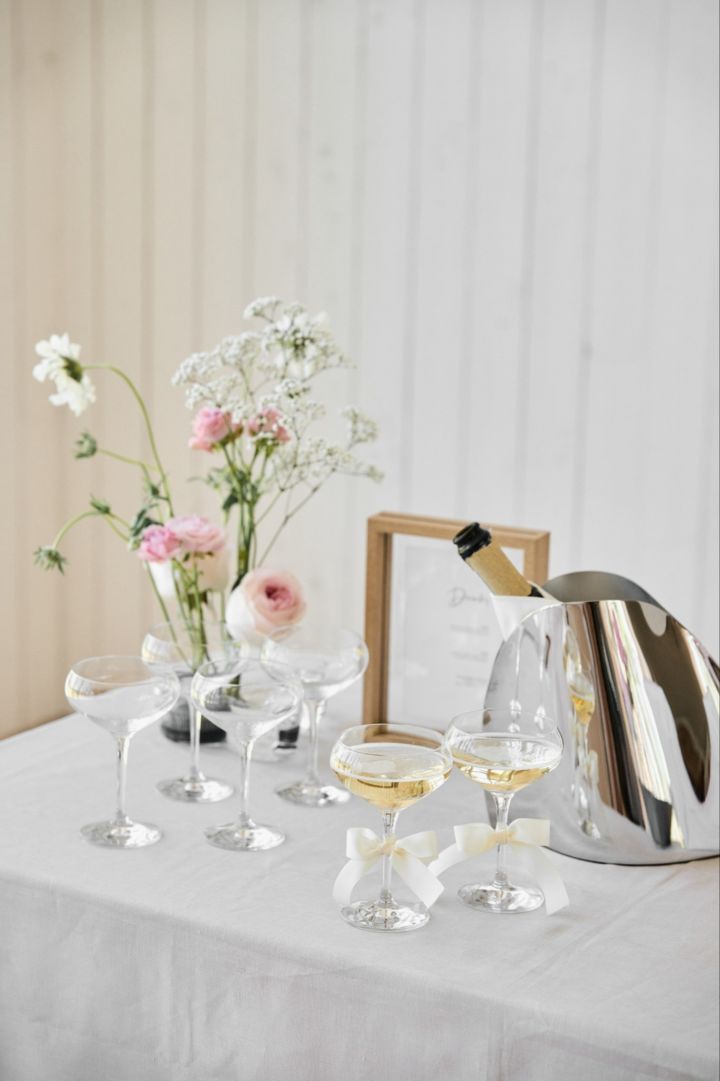 Hier zie je een gedekte drankjes tafel met zes More Coupe glazen van Orrefors en de Indulgence champagne cooler van George Jensen. Op de achtergrond van de tafel zie je een bosbloemen en een ingelijst menukaart.