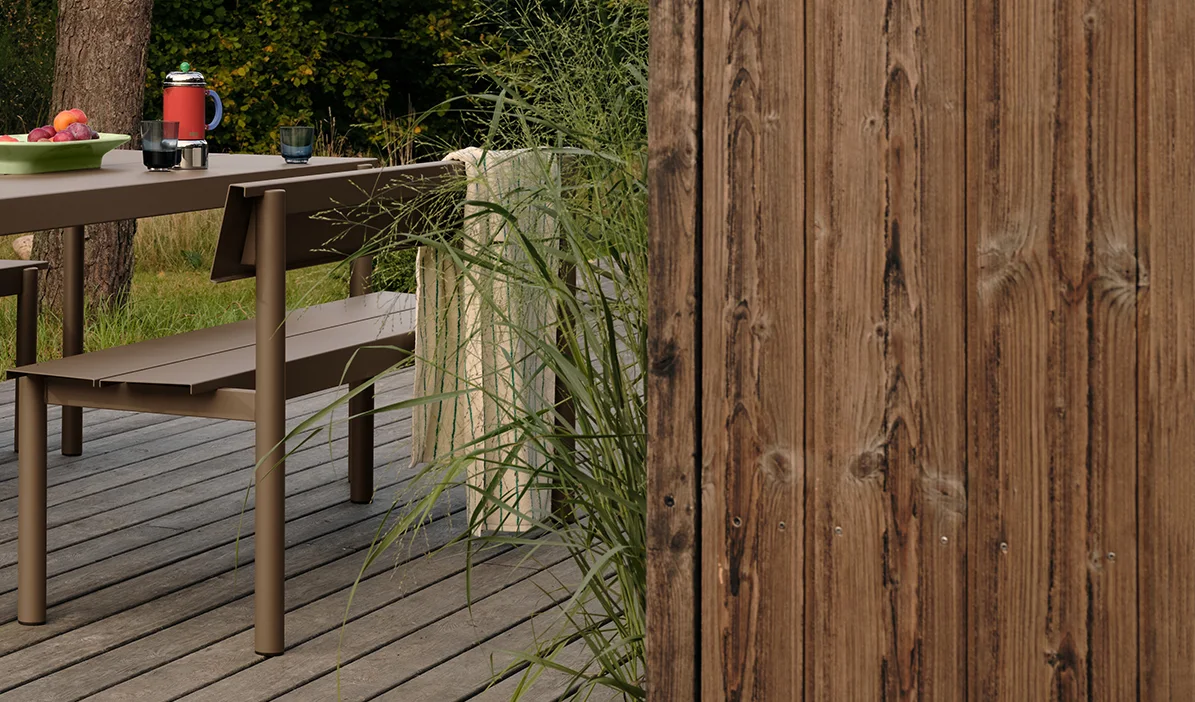 Bruine metalen buitentafel en bank op een houten terras met fruit en drankjes, naast hoog gras en een houten schutting.