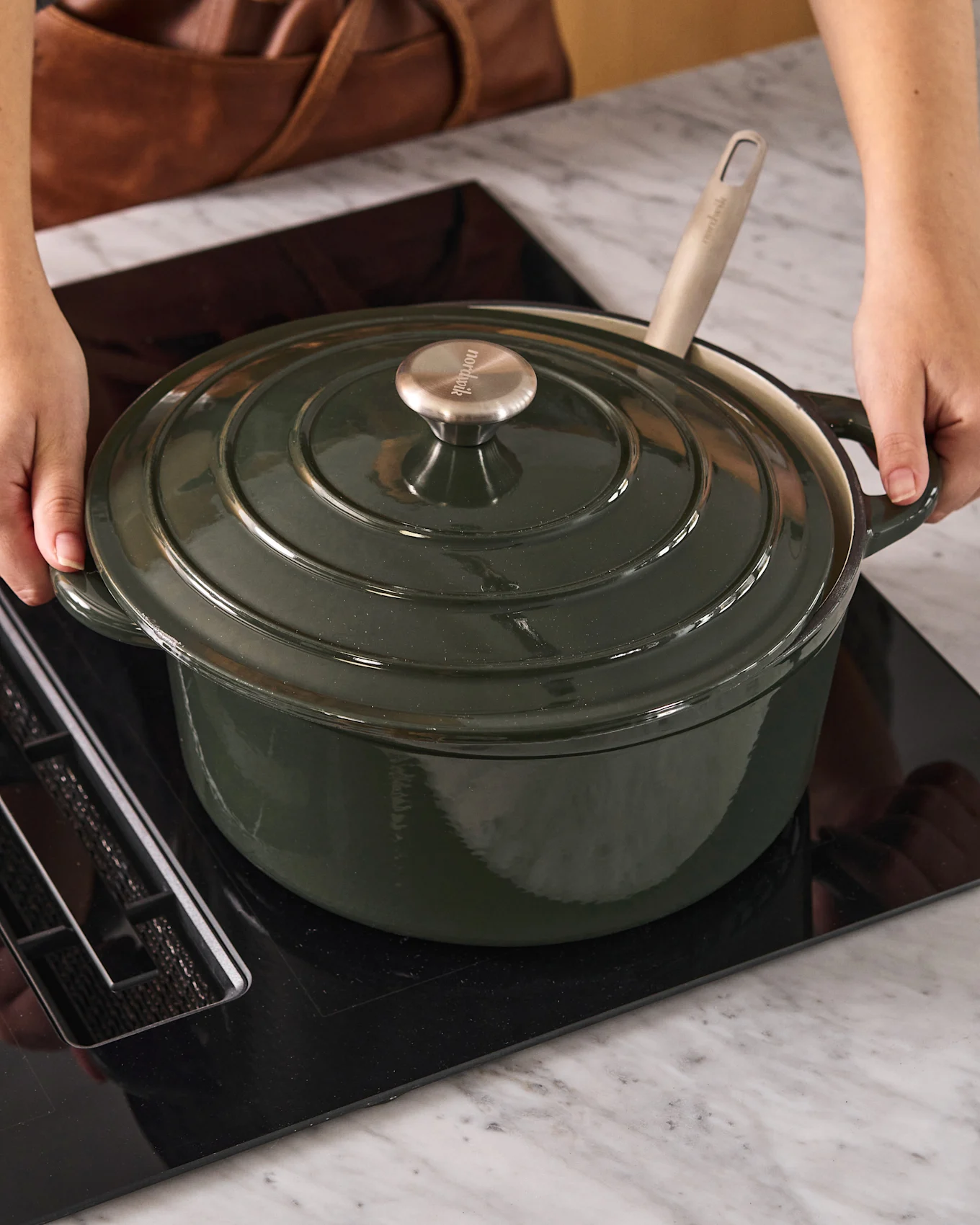 Persoon plaatst een donkergroene braadpan met metalen knop op een zwarte inductiekookplaat.